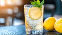 Fresh lemonade cocktail with ice cubes, mint leaf garnish and lemon slice in glass, sparkling drink on blue background with whole citrus fruits.