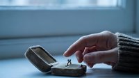 Caucasian woman's hand pointing at vintage engagement ring in antique velvet box on windowsill, moody blue tones evoking nostalgia.