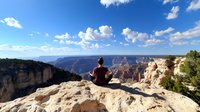 Woman in burgundy shirt sitting in meditation pose on cliff edge overlooking majestic canyon landscape with dramatic rock formations under blue sky with white clouds.