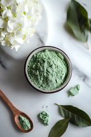 Green clay powder in ceramic bowl with wooden spoon on marble surface, white hydrangea flowers and fresh green leaves create spa wellness composition.