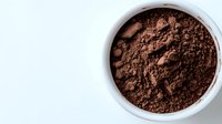 Fine cocoa powder in white ceramic bowl on light background, overhead view showing rich dark brown color and powdery texture for baking and cooking.