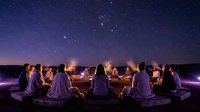 Group of people sitting around campfire under starry night sky enjoying outdoor gathering and bonding experience together in wilderness setting.