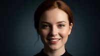 Young professional woman with red hair smiling confidently against dark background. Corporate headshot for business presentations and marketing materials.