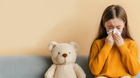 Young Asian woman in yellow sweater blowing nose with tissue paper while sitting on couch next to plush teddy bear, showing cold and flu symptoms on beige background.