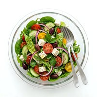 Fresh garden salad with tomatoes, cucumbers, feta cheese and mixed greens in glass bowl with fork and knife on white background, ready for healthy eating.