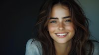 Young Caucasian woman with bright smile, blue eyes and natural brown wavy hair wearing grey sweater against dark background, radiating genuine happiness and confidence.