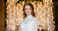 Young Caucasian woman in elegant white lace dress smiling at camera with warm golden bokeh lights background for holiday celebrations and events.