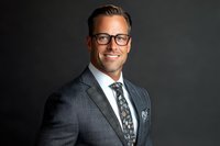 Confident male business professional in dark suit and glasses smiling against dramatic black background, representing success and leadership in corporate setting.