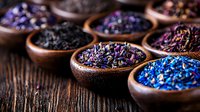 Colorful dried flower petals and tea leaves in wooden bowls arranged on rustic table, selective focus on vibrant purple and blue botanical ingredients.