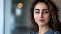 Young Mediterranean woman with long dark hair and natural makeup smiling confidently at camera. Professional headshot with soft bokeh background in office setting.