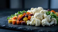 Fresh chopped vegetables including cauliflower florets, diced carrots, and green herbs arranged on dark slate surface for healthy cooking preparation.