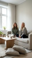 Two young Caucasian women friends relaxing together on beige sofa in bright minimalist living room with plants and natural light for lifestyle content.