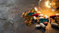 Colorful pharmaceutical capsules and pills scattered on dark surface with glowing light bulb representing medical innovation and healthcare breakthrough concepts.