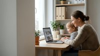 Young mother working from home office with baby on lap using laptop computer at wooden desk near window with plants and organized shelves.