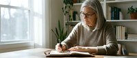 Senior woman with gray hair and glasses writing in notebook at wooden desk by window, surrounded by bookshelves and potted plants in bright home office setting.