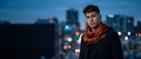 Young handsome Caucasian man in stylish scarf standing on urban rooftop at dusk with blurred city lights background for lifestyle and fashion content.