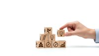 Businessman hand arranging wooden blocks with business icons including growth charts, gears, and success symbols on white background for corporate strategy.
