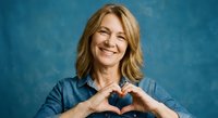 Middle-aged Caucasian woman making heart shape with hands, smiling warmly in denim shirt against blue background for wellness and self-care campaigns.