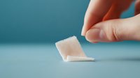 Small piece of white medical gauze bandage held by fingers against blurred turquoise background, demonstrating sterile first aid material in healthcare setting.