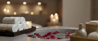 Luxury spa setting with rolled white towels, red rose petals, candles and warm ambient lighting creating relaxing wellness atmosphere for massage therapy.