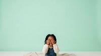 Young South Asian male child with hands covering face in gesture of stress, frustration or fatigue against soft mint green background, suitable for educational materials, parenting content, mental health awareness.