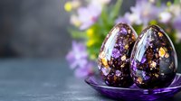 Dark chocolate Easter eggs decorated with purple edible glitter, gold flakes and dried flowers on purple glass plate against blurred floral background.
