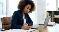 Young African American businesswoman writing notes while working on laptop in modern office environment for professional business content.
