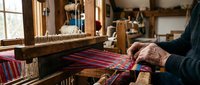 Elderly craftsman weaving colorful striped textile on traditional wooden loom in rustic workshop. Handmade fabric production, artisan skills, heritage crafts.