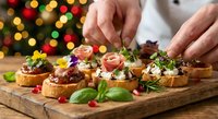 Chef hands garnishing gourmet bruschetta appetizers on wooden board with festive Christmas lights creating warm holiday dining atmosphere.