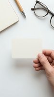 Hand holding blank white business card with notebook, pen and glasses on clean desk workspace for corporate branding and professional identity design.