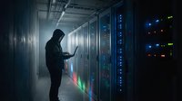 Hooded figure working on laptop in modern data center with glowing server racks and colorful LED lights creating atmospheric cybersecurity environment.