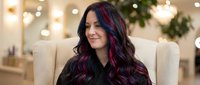 Woman with long dark hair featuring vibrant purple and magenta highlights, seated in a cream-colored chair in an elegantly decorated interior with festive lighting and greenery. For beauty, fashion, and lifestyle content.