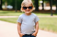 Cheerful young child wearing sunglasses and casual grey t-shirt standing outdoors in park with hands in pockets, expressing confidence and joy on sunny summer day.