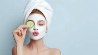 Young woman with facial clay mask holding cucumber slice over eye, hair wrapped in towel against light blue background. Skincare and beauty routine concept.