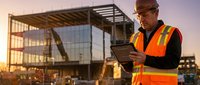 Middle-aged Caucasian construction foreman in safety vest using tablet at modern building site during golden hour sunset for project management and planning.