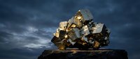 Golden metallic pyrite mineral specimen with cubic crystals on dark stone base against dramatic stormy sky background for geology education and collection display.
