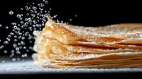 Stack of thin delicate crepes or pancakes with powdered sugar and water drops splashing in dramatic lighting on dark background, macro food photography.