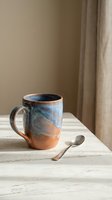 Handmade ceramic mug with blue and terracotta glaze on wooden table with spoon in warm natural light for cozy morning coffee or tea rituals.