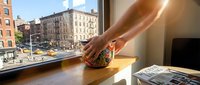 Caucasian woman's hand holding decorative globe near sunny window with urban cityscape view, workspace setting.