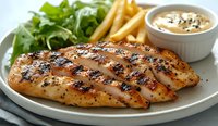 Grilled chicken breast with black pepper seasoning served on white plate alongside crispy french fries, fresh green salad and creamy dipping sauce.