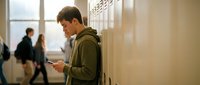Young Caucasian male student using smartphone while standing by school lockers in hallway during break time with classmates walking in background.