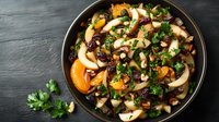 Fresh fruit and nut salad with apples, mandarin oranges, cranberries, and parsley garnish served in dark bowl on black stone background.