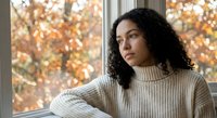 Young mixed-race woman in cozy sweater looking thoughtfully out window during autumn season with warm natural lighting for lifestyle content.