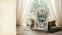 Ceramic mug with botanical leaf pattern sits on rustic wooden table near window with natural lighting for cozy home decor and morning coffee rituals.