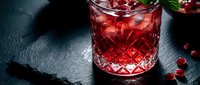 Elegant ruby red pomegranate cocktail in crystal glass with ice, fresh mint, and scattered seeds on dark slate surface.