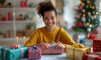 Young African American woman with curly hair in yellow sweater wrapping Christmas presents at home, smiling while decorating colorful gift boxes near decorated tree.