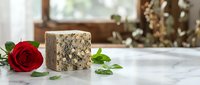Aged blue cheese block with crumbly texture displayed on marble surface alongside fresh red rose and green herbs in bright kitchen setting.