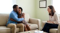 African American couple embracing on beige sofa during therapy session with female counselor in bright office setting for relationship support.