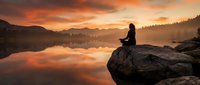 Woman in silhouette meditating on rocky outcrop overlooking serene lake at sunrise, surrounded by misty mountains and forested landscape, conveying peace and contemplation.