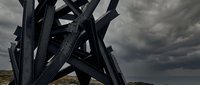Abstract black metal sculpture with angular geometric forms against dramatic stormy sky. Modern art installation with intersecting beams and industrial design elements.